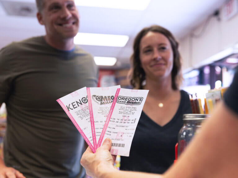 In the foreground, an arm holds out several Lottery draw games tickets over a counter. In the background, a man and woman, customers, smile on the other side of the counter